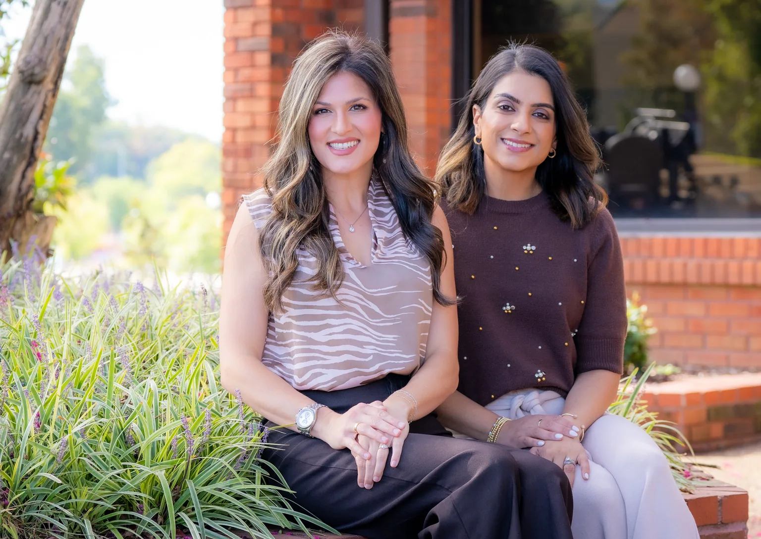 Julia Klug, PA-C and Priya Patel, PA-C sitting together on a brick planter outside their office